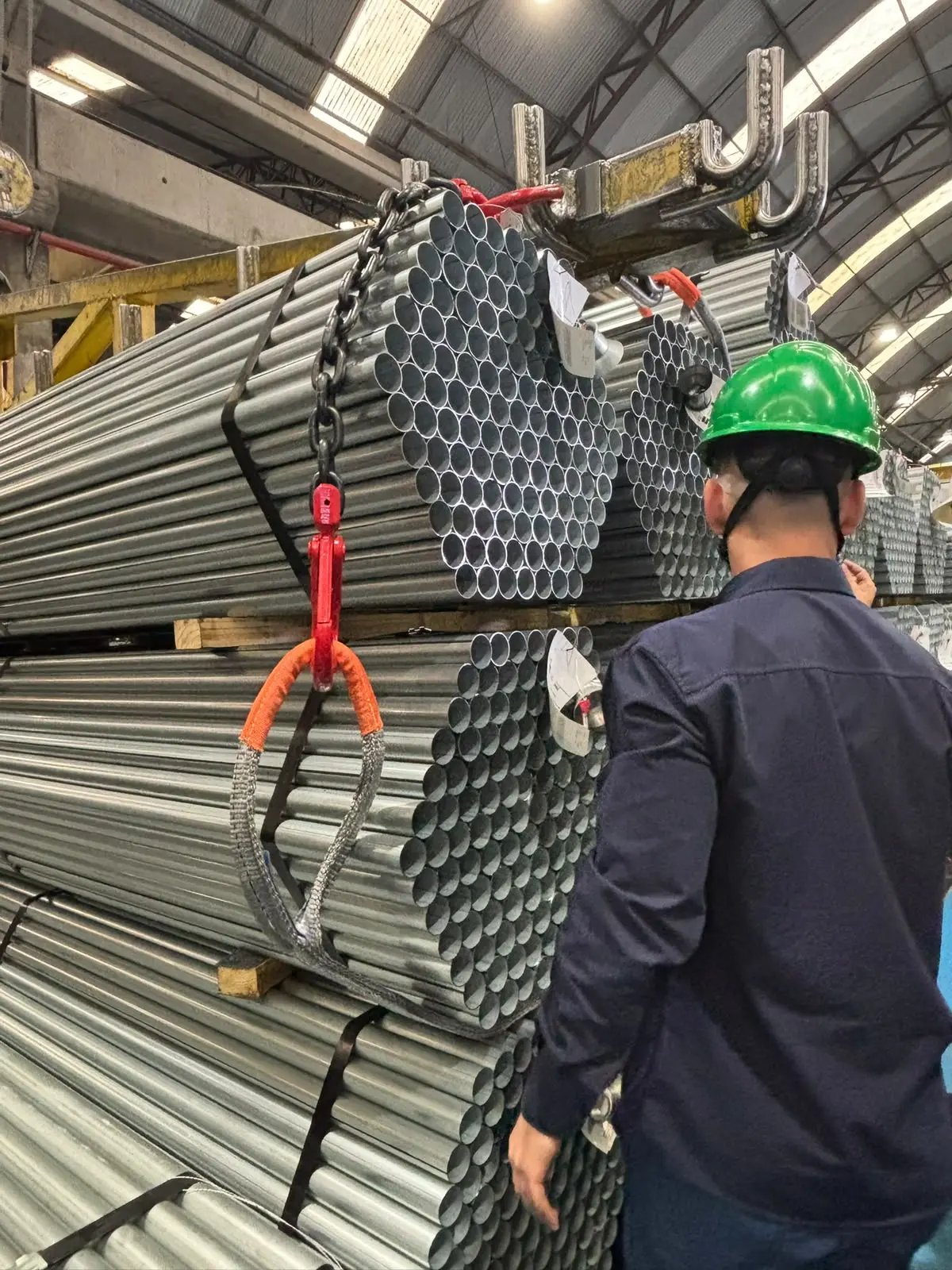 Profissional da Qualimax com capacete acompanhando a movimentação de feixes de tubos metálicos com cinta de elevação